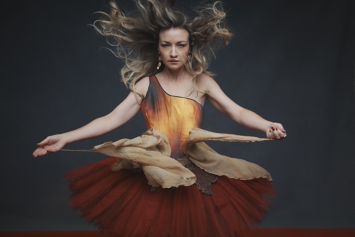 Australian singer Kate Miller-Heidke posing in an orange and red dress with windblown blonde hair against a dark backdrop.